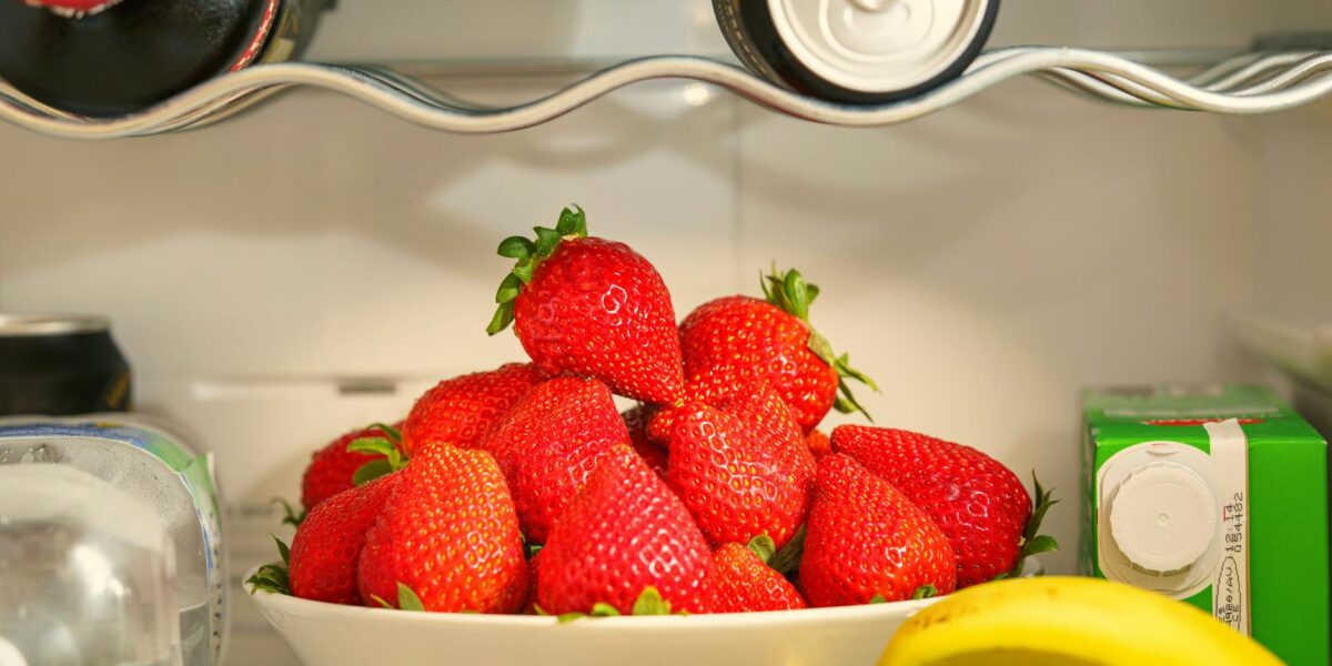 A bowl of ripe strawberries in a refrigerator, surrounded by drinks and bananas, showcasing vibrant freshness.
