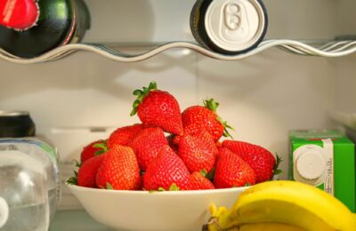 A bowl of ripe strawberries in a refrigerator, surrounded by drinks and bananas, showcasing vibrant freshness.