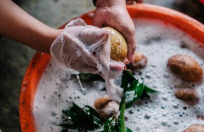 Hands washing potatoes in soapy water with plant leaves using a white cloth.