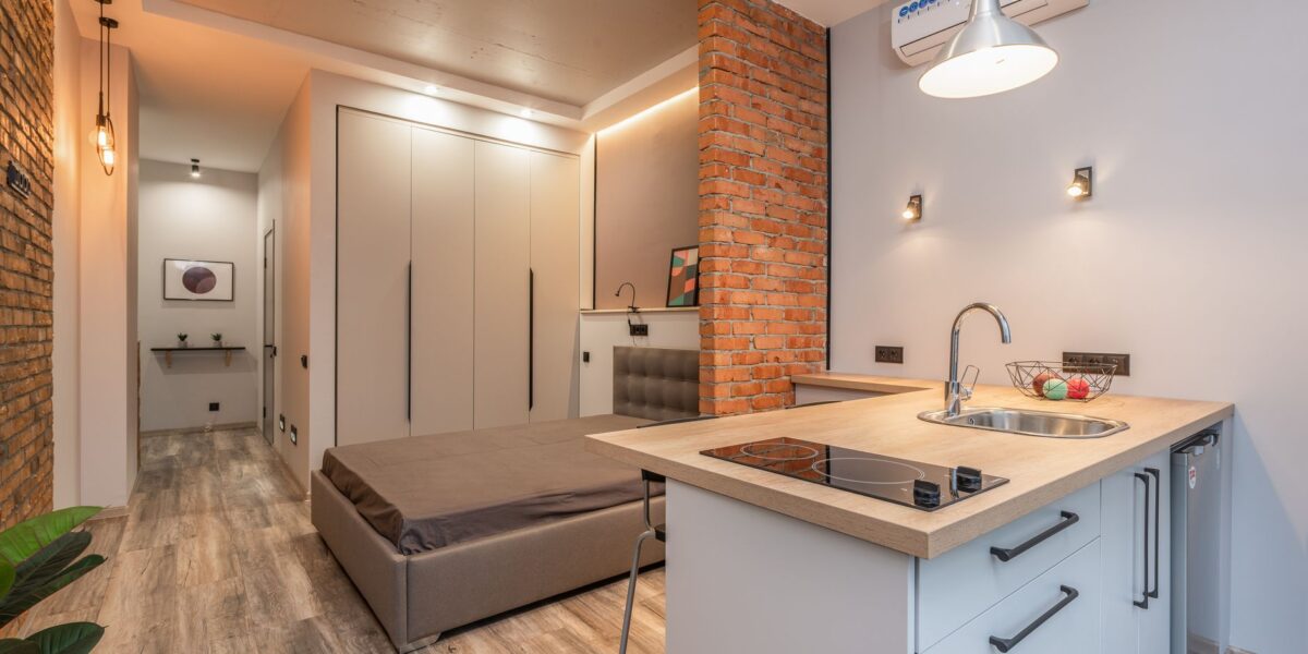 Miniature kitchen with modern appliances and bedroom behind brick partition in small loft style apartment