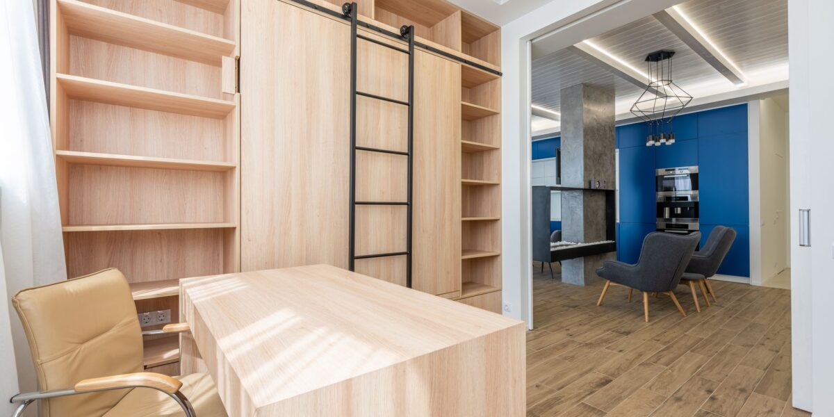 Wooden table and armchair placed near bookcase in office zone and stylish furniture in kitchen