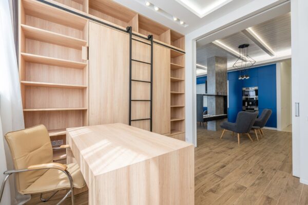 Wooden table and armchair placed near bookcase in office zone and stylish furniture in kitchen