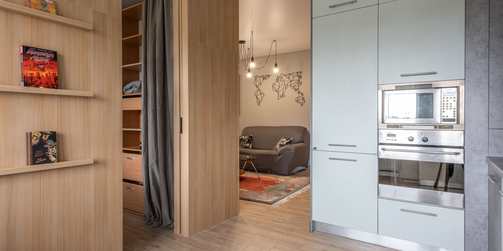 Modern apartment with cabinets with oven and microwave in kitchen near wooden shelves with books near entrance in room with wardrobe with curtains near living room with couch and carpet