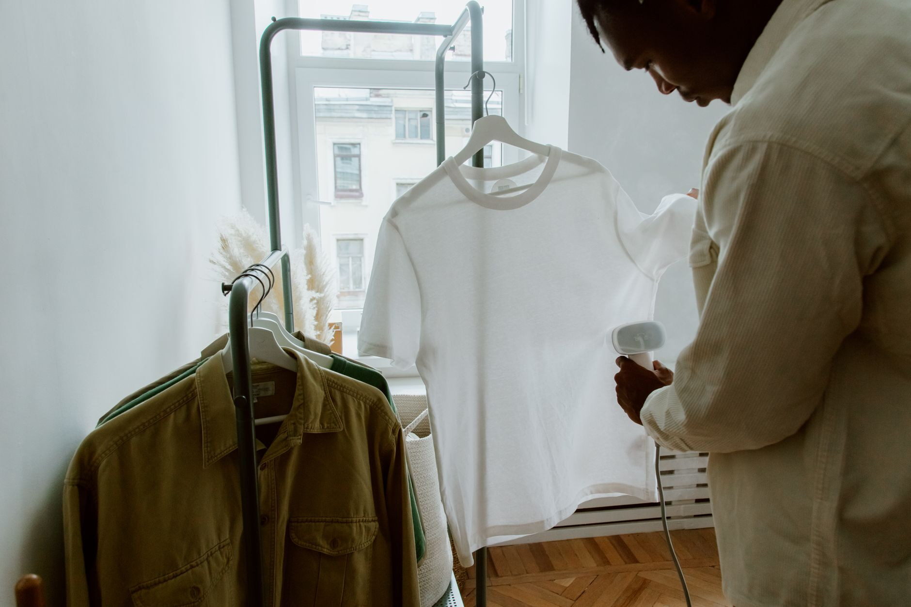 A man uses a steam iron on a white shirt hanging from a clothing rack indoors.