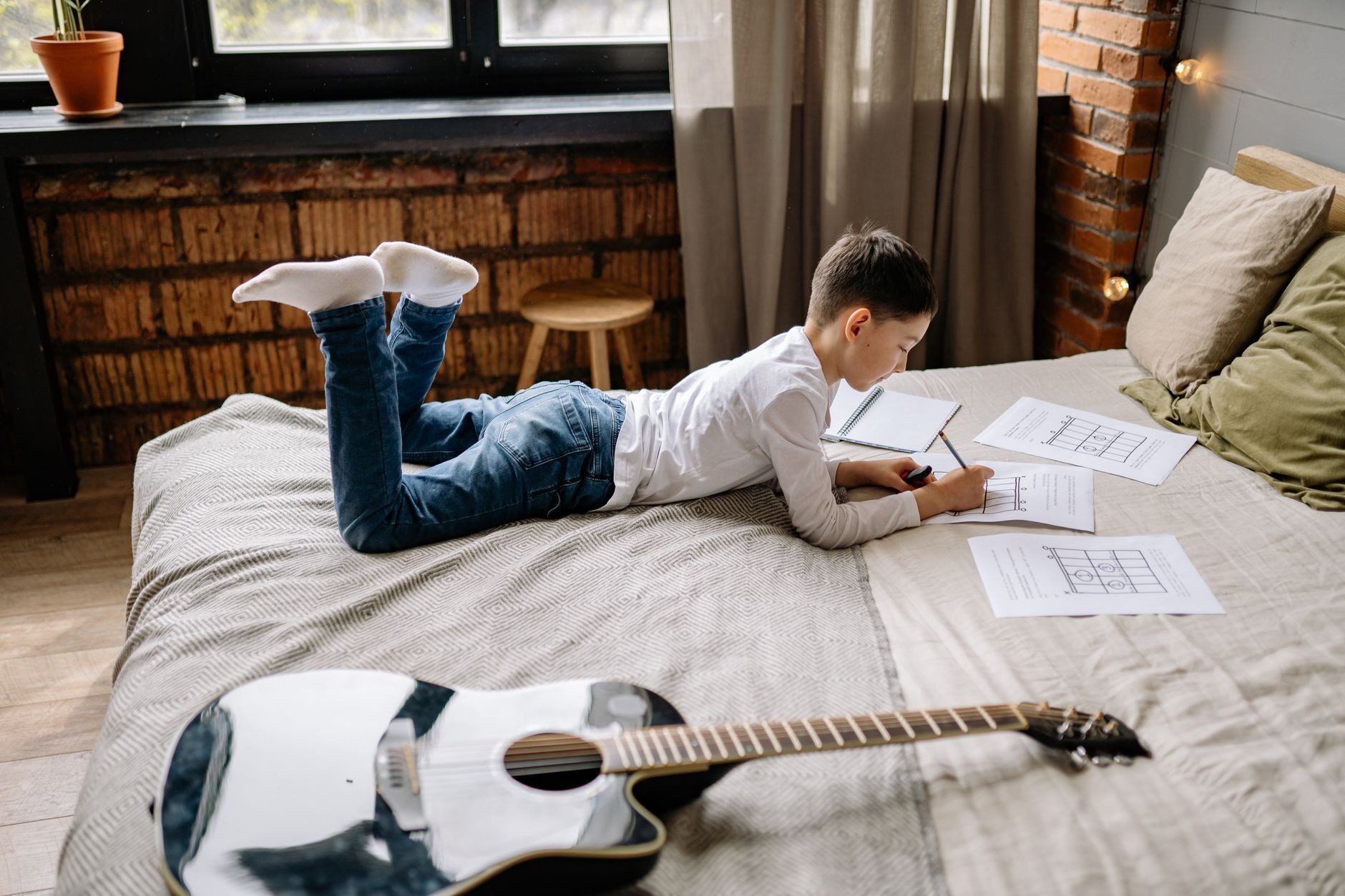 A young boy practicing guitar chords on paper while lying on a bed in a cozy room.